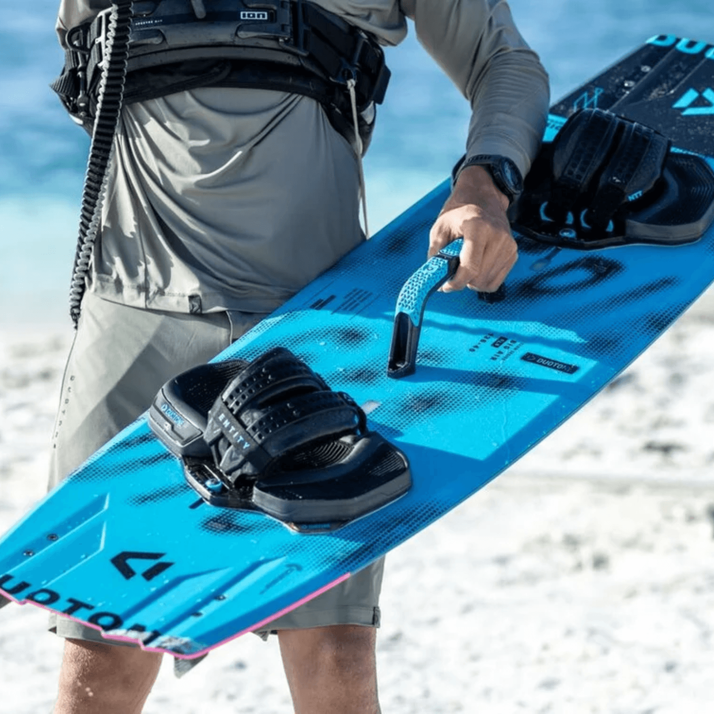 duotone-big-air-handle-alt2 Rider carrying a Duotone twintip kiteboard by the Big Air Handle, with Entity pads and straps and the WOO sensor mount visible on the grip.